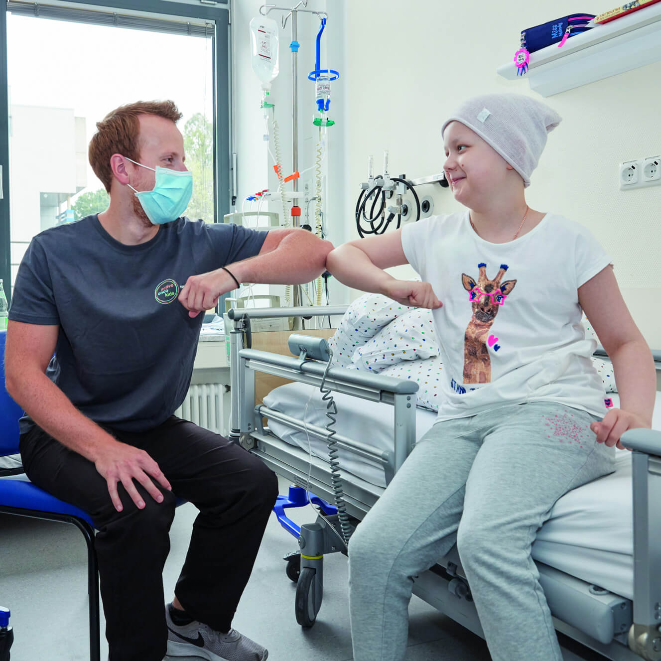 Ein Sporttherapeut und ein krebskrankes Mädchen wie sie im Patientenzimmer sitzen und einen Elbogencheck geben