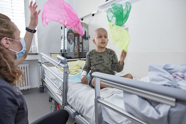 Eine Sporttherapeuthin die gemeinsam mit einem krebskranken Jungen, der im Patientenbett liegt, spielerisch Bewegungen auffordert. Sie wirft bunte Tücher in die Luft und der Junge muss diese fangen.