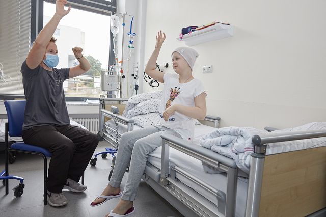 Ein Sporttherapeut der gemeinsam mit einer krebskranken Jugendliche leichte Körperübungen im Patientenzimmer durchführt